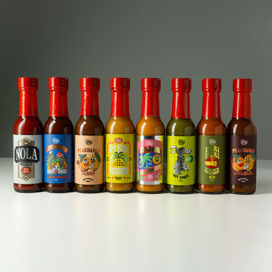 Eight small hot sauce bottles lined up on a white surface against a dark gray gradient background. The bottles have red caps and colorful labels with different designs and text, including NOLA, Peachango Mango, Pineappleachiachi, Tequila, Dollar, and Peachango Habanero.