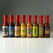 Eight small hot sauce bottles lined up on a white surface against a dark gray gradient background. The bottles have red caps and colorful labels with different designs and text, including NOLA, Peachango Mango, Pineappleachiachi, Tequila, Dollar, and Peachango Habanero.